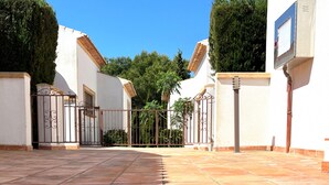 A courtyard with a tiled floor, a metal gate, and a building with a white exterior and a brown roof. - 0356 Monte Javea Calicanto (Javea / Xàbia)