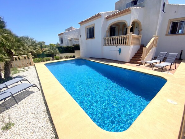 A rectangular swimming pool with clear blue water, surrounded by a light-colored deck. There are two lounge chairs on the right side of the pool.