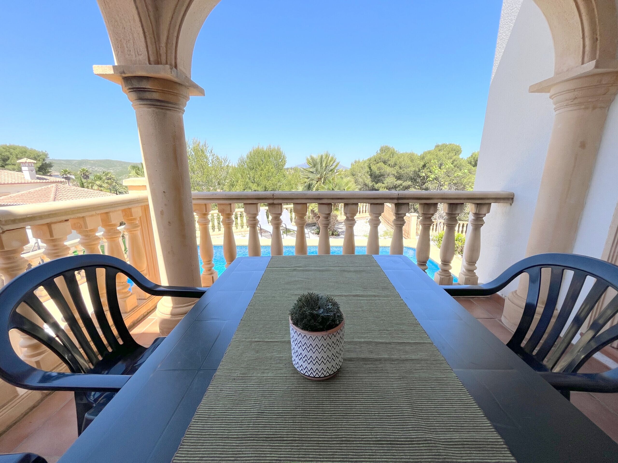 A balcony with a table and chairs, a potted plant, and a view of a swimming pool and greenery.
