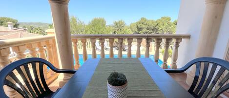 A balcony with a table and chairs, a potted plant, and a view of a swimming pool and greenery.