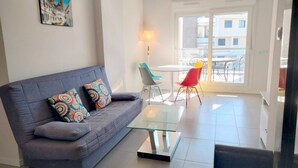 Un salon moderne avec un canapé gris, des coussins colorés, une table basse en verre et une vue sur le balcon extérieur.