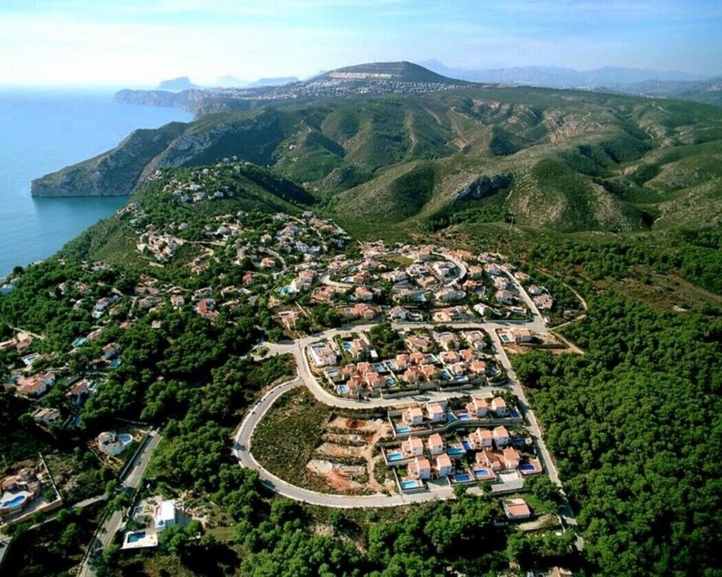 An aerial view of a coastal residential area with houses arranged in a semi-circular pattern, surrounded by lush greenery and overlooking the sea.