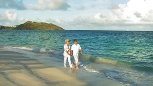 Una playa con dos personas caminando por la orilla, una isla distante y un velero en el agua.