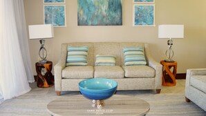 Un salón con un sofá color beige, dos mesitas de noche con lámparas, un cuenco azul sobre una mesa de centro y tres cuadros abstractos en la pared.