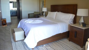Habitación de hotel con una cama grande, una mesita de noche de madera, una mesita junto a la cama con una lámpara, una silla y vistas al baño.