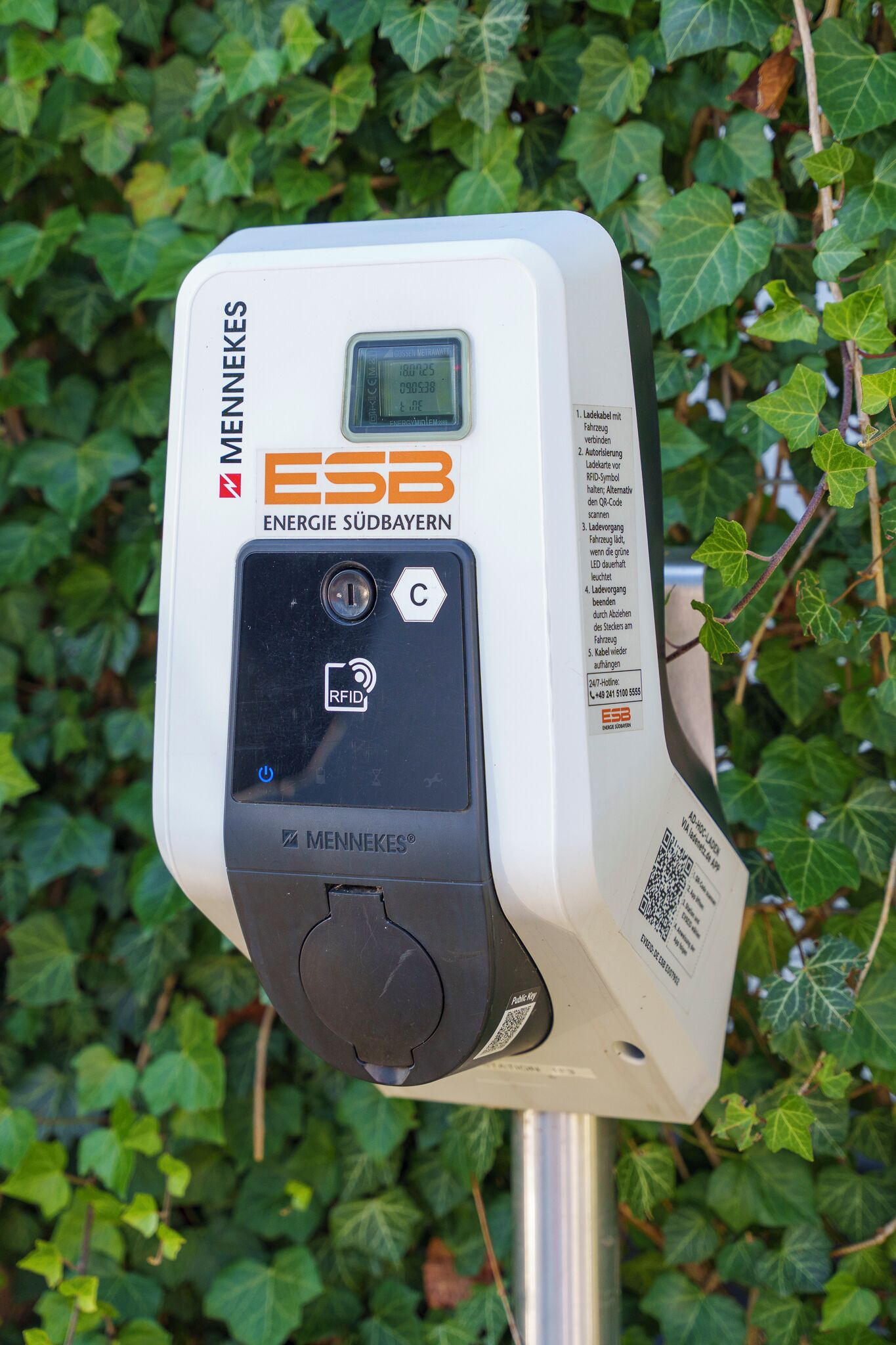 Ein MENNEKES Energiemessgerät mit digitaler Anzeige, RFID-Lesegerät und einem QR-Code, vor einem Hintergrund aus grünen Efeu-Blättern.
