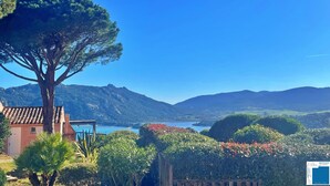 A coastal landscape with a large tree, a building with a tiled roof, and a view of the sea and mountains.