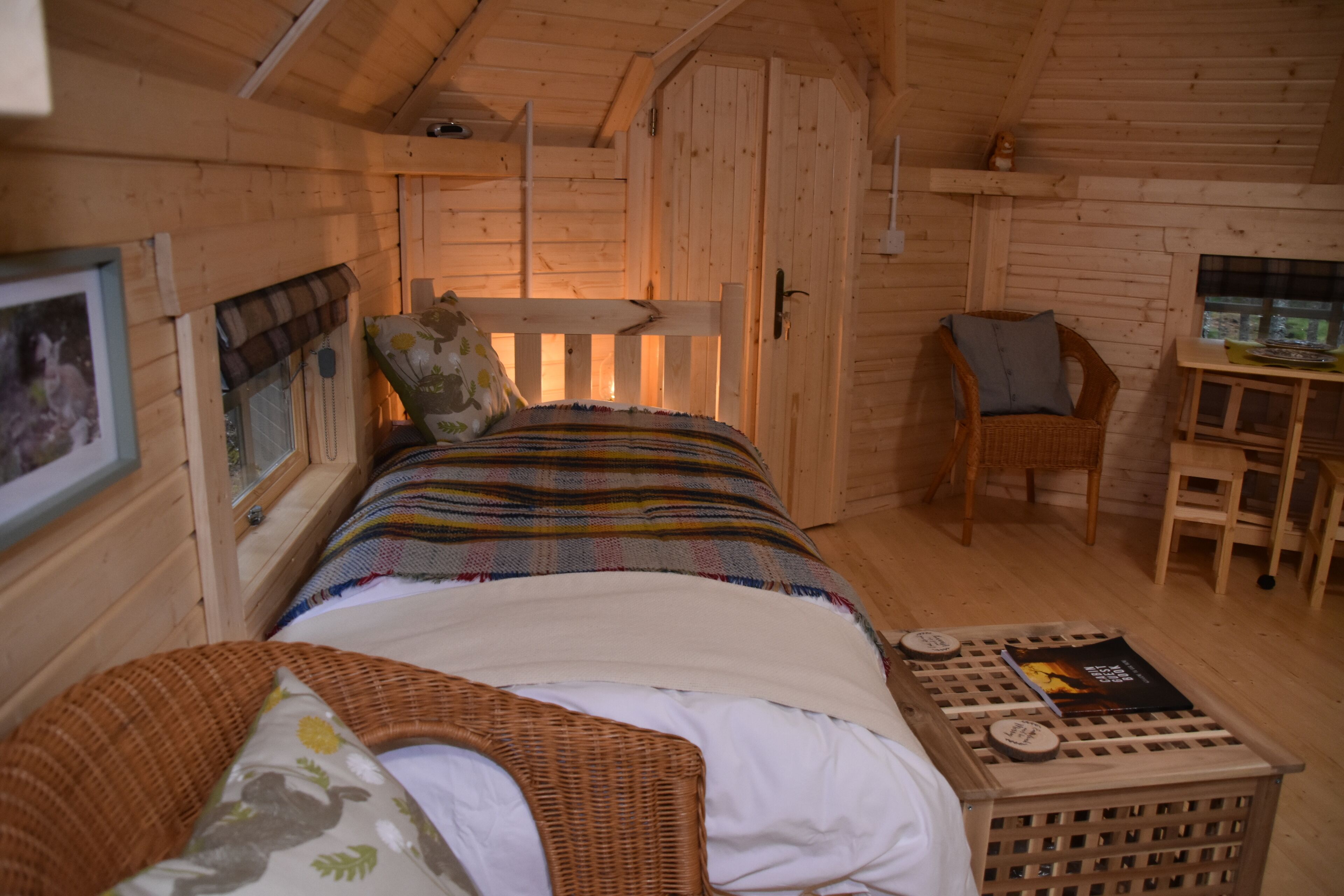 A cozy wooden cabin room with a bed, a chair, a small table, and a framed picture on the wall.