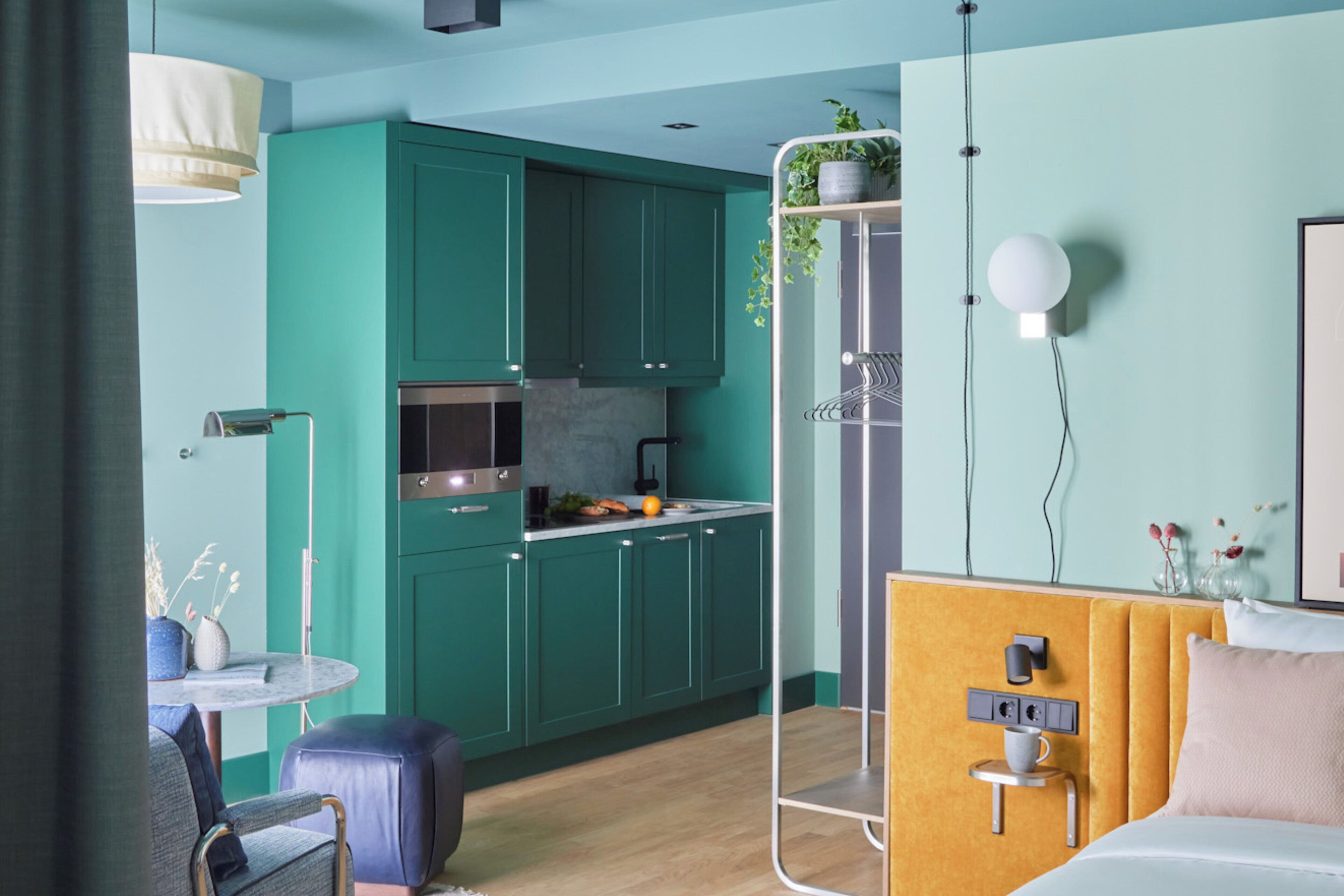 A modern hotel room with a bed, bedside table, and a view of the kitchen.