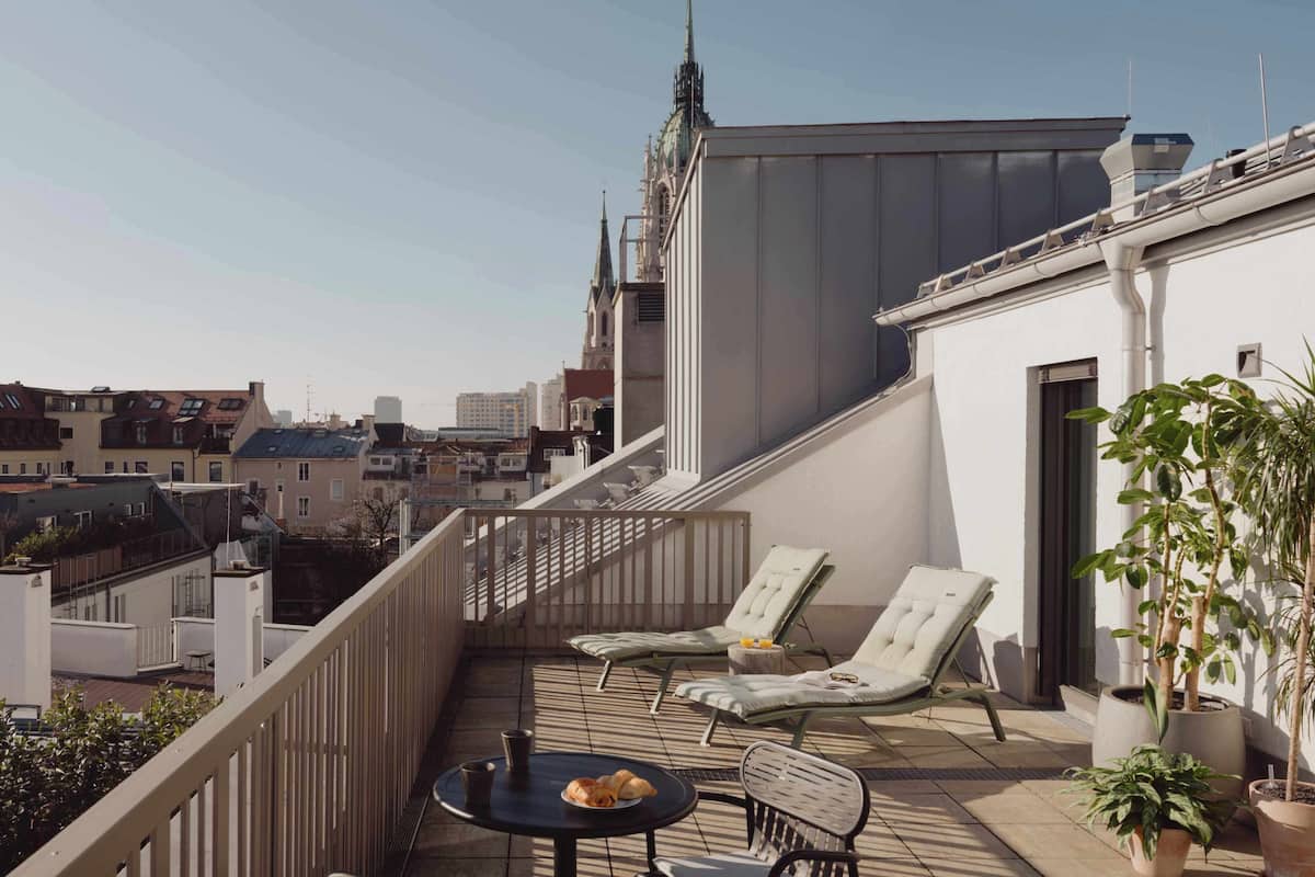 A rooftop terrace with two lounge chairs, a small table with a cup and pastries, and potted plants.