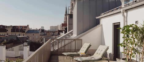 A rooftop terrace with two lounge chairs, a small table with a cup and pastries, and potted plants.