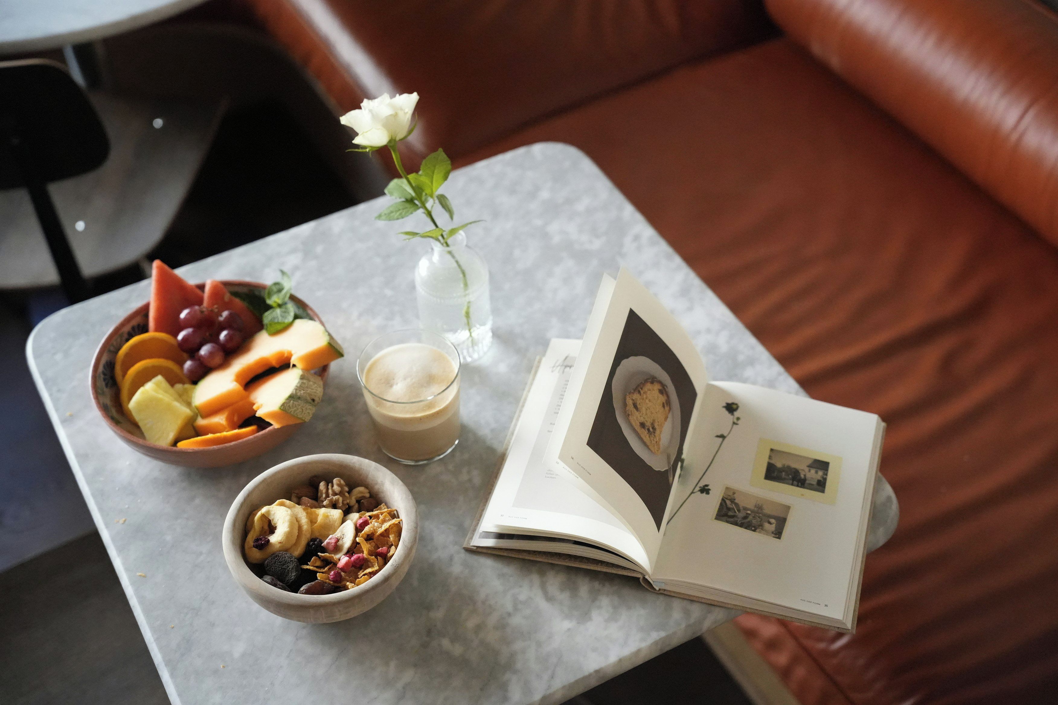 A table with a bowl of fruit, a glass of beverage, a bowl of cereal, a vase with a flower, and an open book with photos.