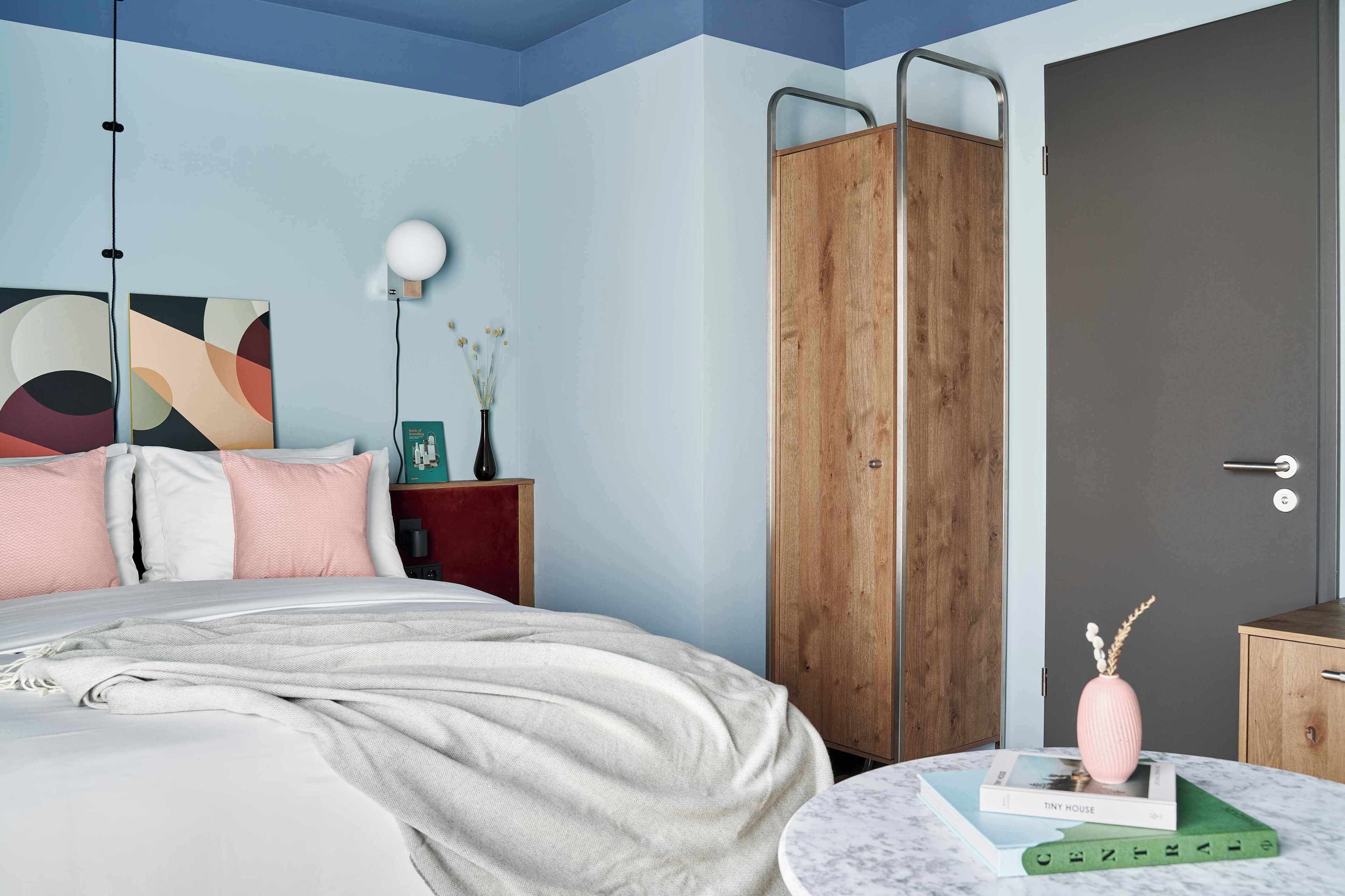 A bedroom with a bed, a wardrobe, a round table with books, a lamp, and a wall art piece.