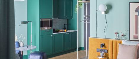 A compact room with teal cabinetry, a kitchenette, a bed with a headboard, and a small table with a chair.