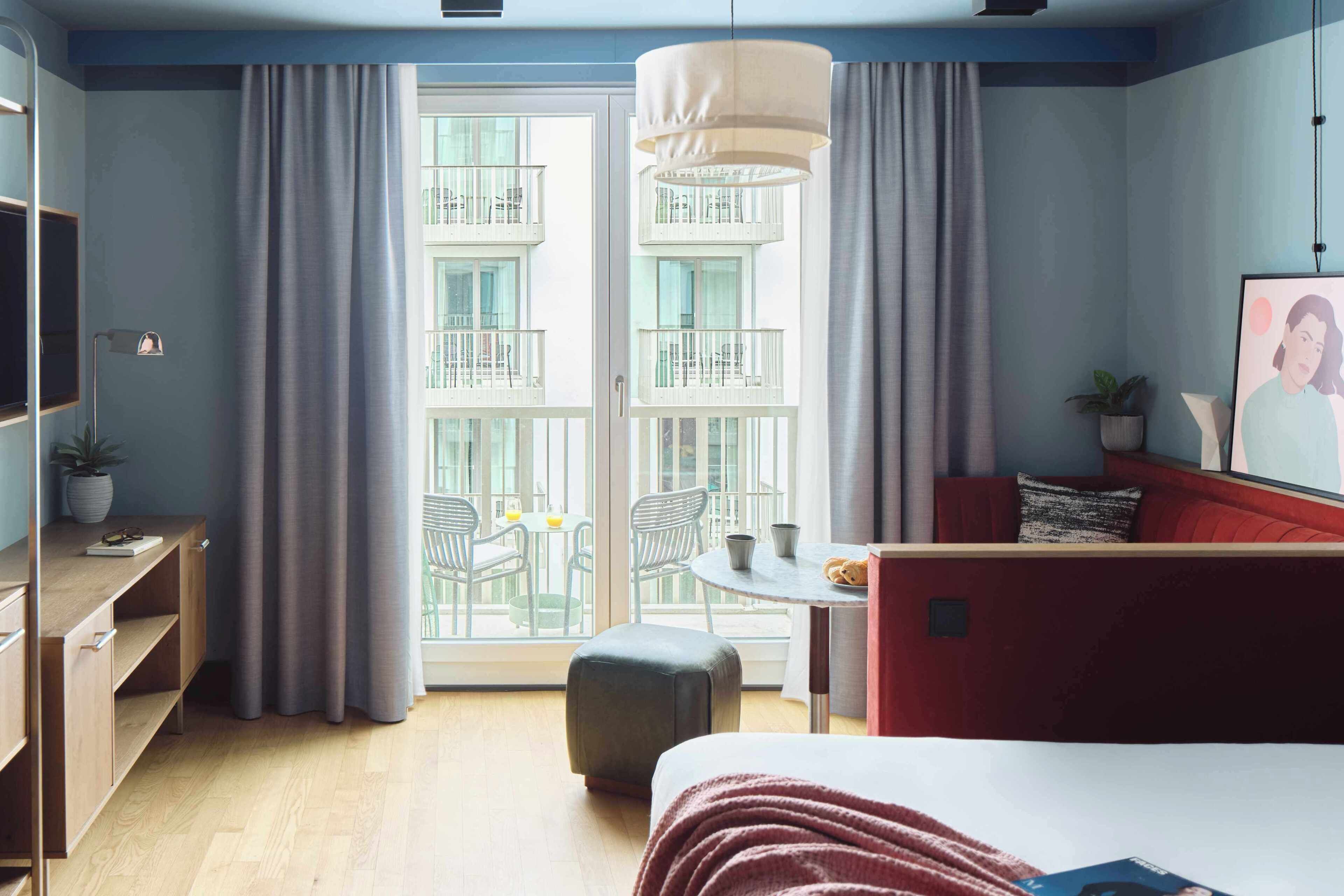Hotellihuone, jossa on sänky, työpöytä, tuoli, pieni pöytä, punainen sohva ja parveke, jolta on näkymä rakennuksen parvekkeille.