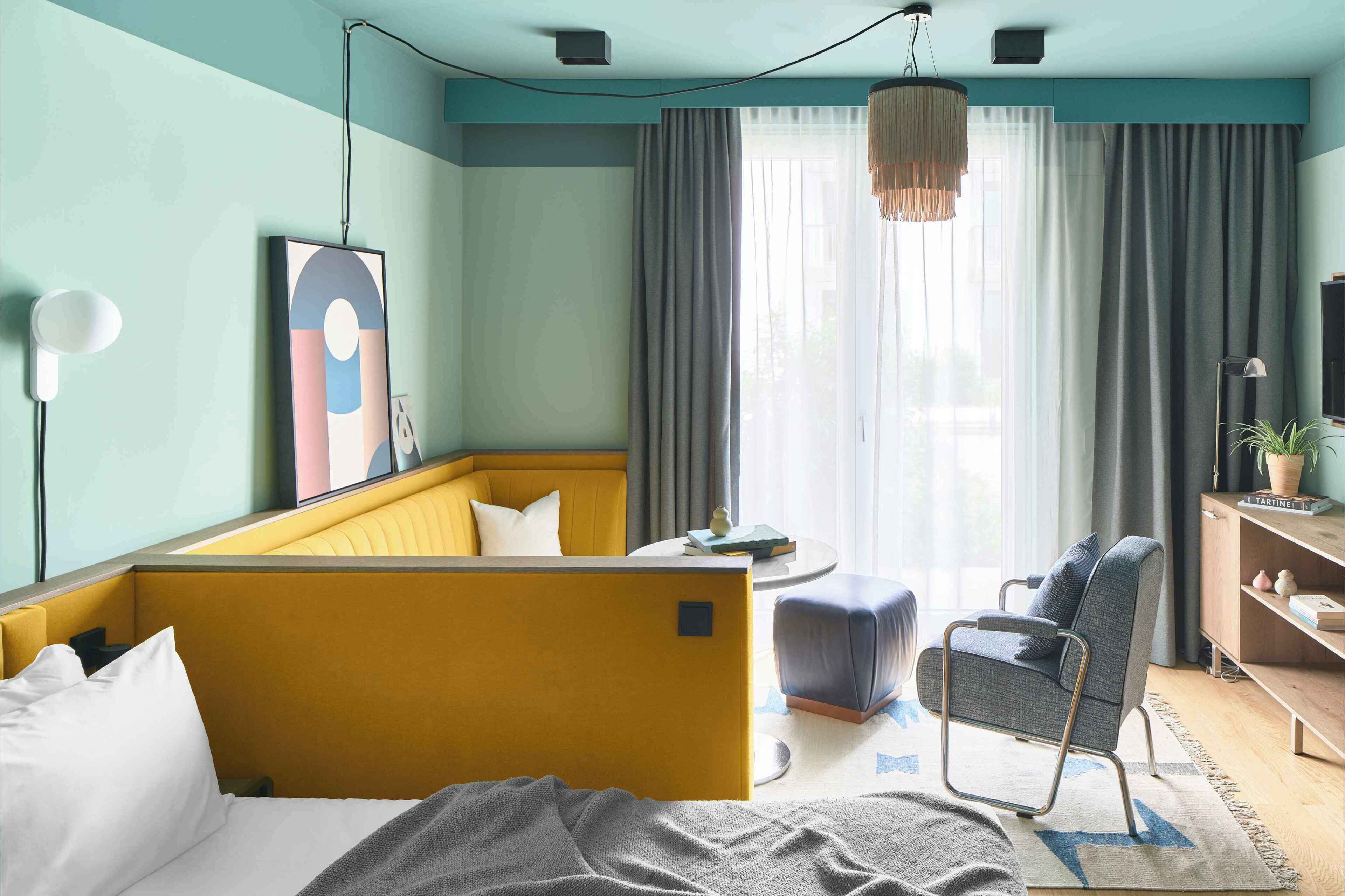 A modern hotel room with a yellow sofa, a grey armchair, a small round table, and a bookshelf.