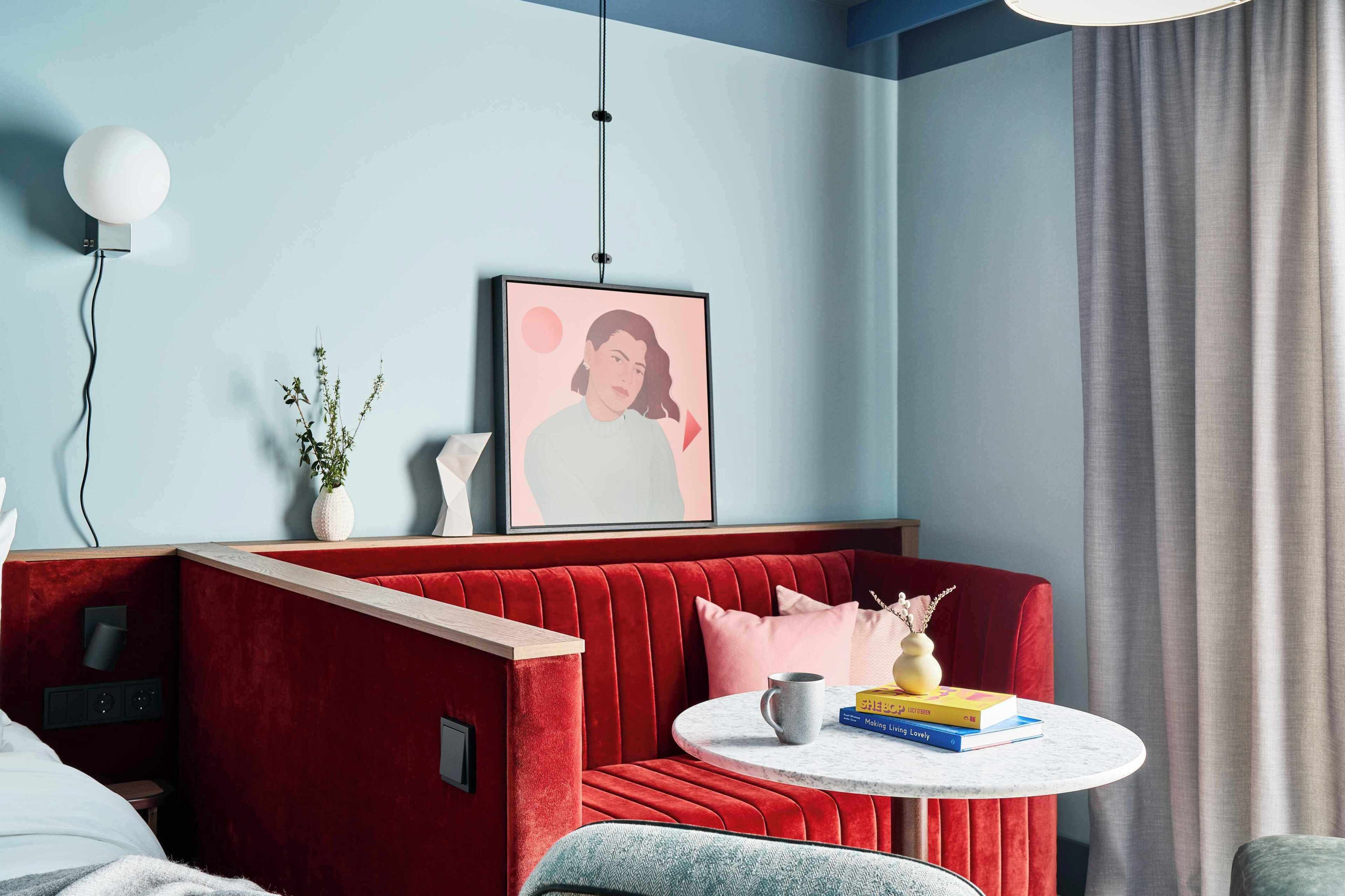 Quarto moderno com sofá de veludo vermelho, uma mesa redonda com livros, uma obra de arte emoldurada na parede e uma planta em vaso.
