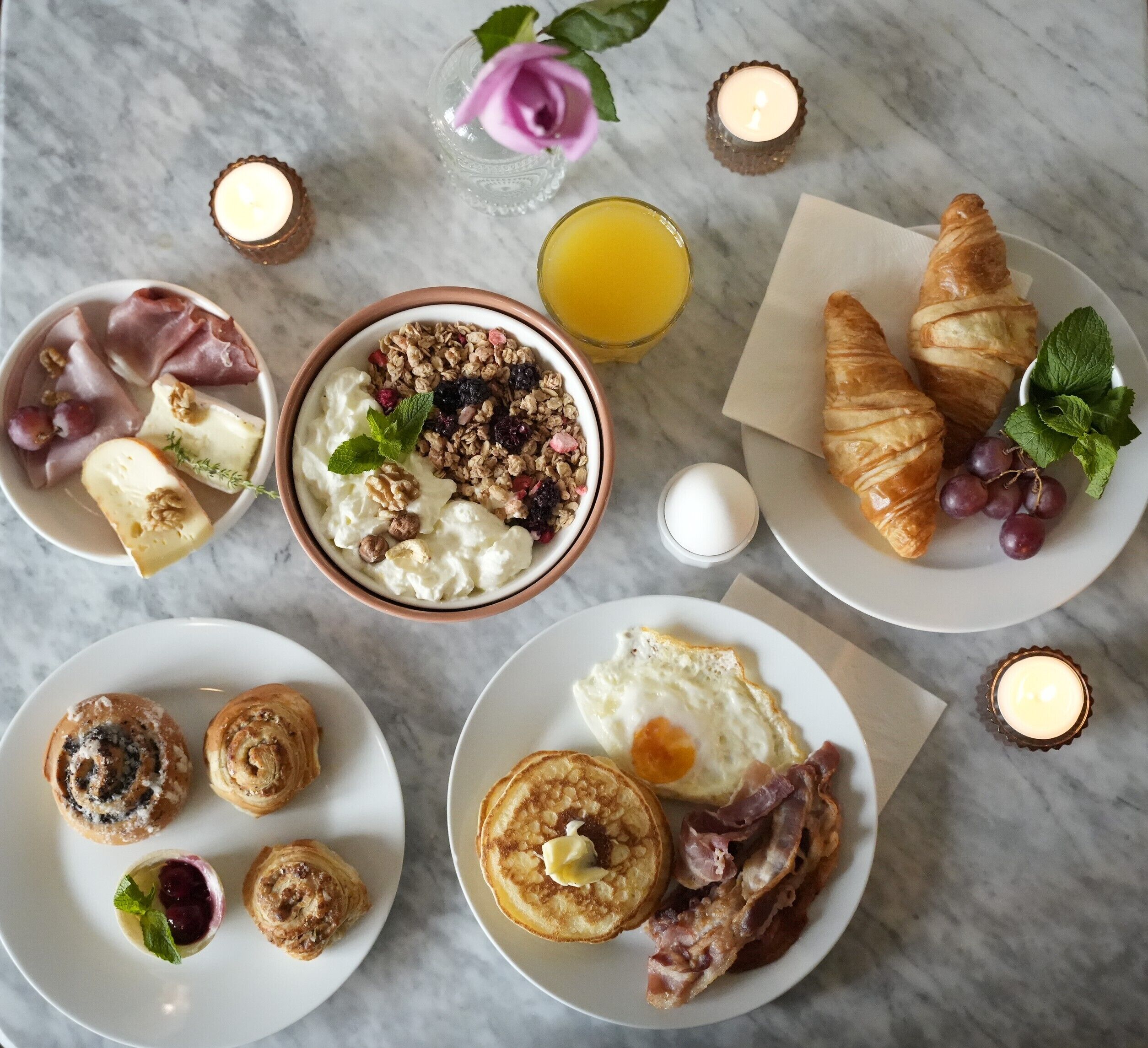 Une table mise pour le petit-déjeuner, avec différents aliments disposés sur des assiettes, une bougie et une rose.