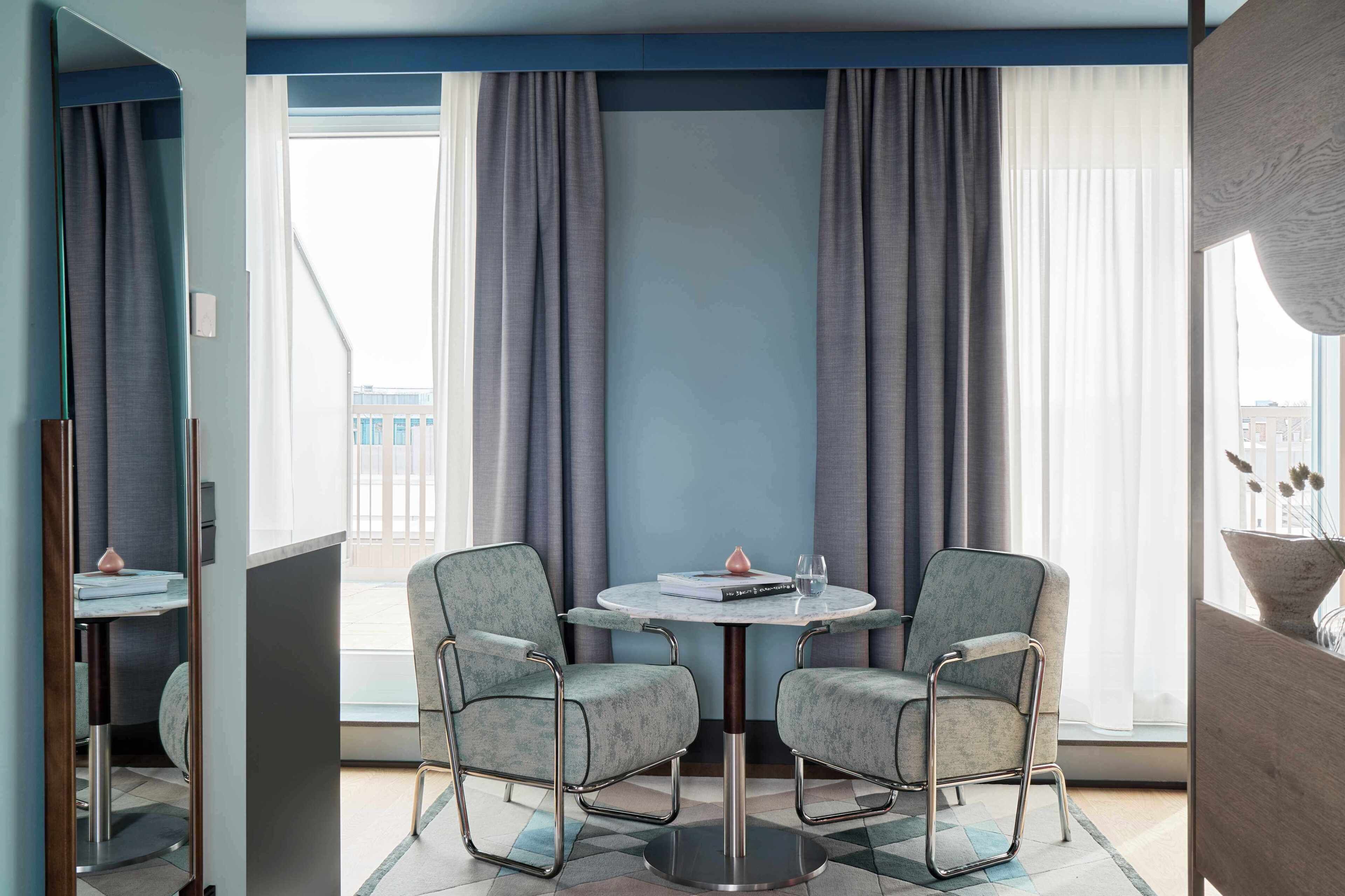 Una habitación de hotel moderna con una mesa redonda, dos sillas, un ventanal con cortinas y vistas a edificios.
