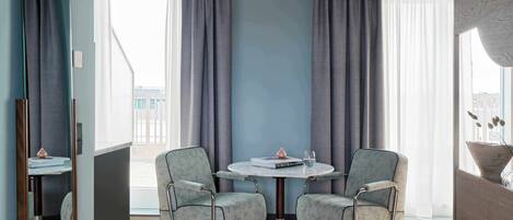A modern hotel room with a round table, two chairs, a large window with curtains, and a view of buildings outside.