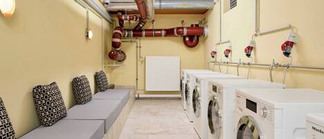 A laundry room with multiple washing machines, a bench with patterned cushions, and visible piping on the walls.