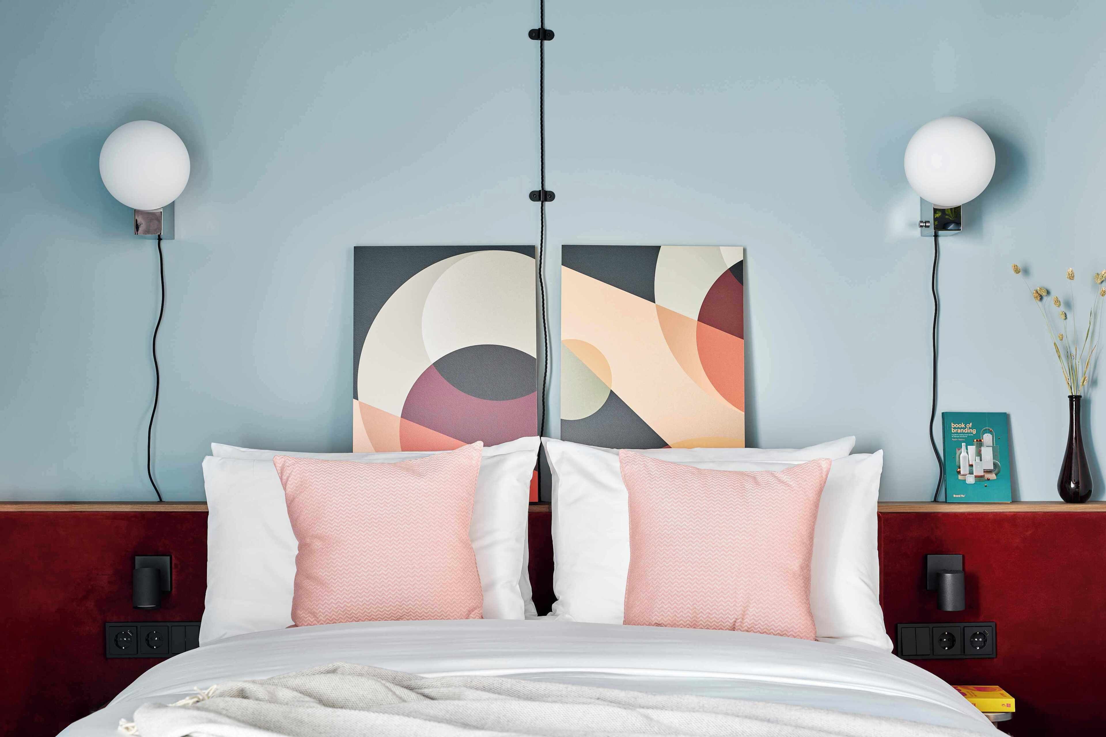 Una habitación de hotel con una cama, dos lámparas fijadas a la pared, un cuadro, un florero con flores y un libro en la mesita de noche.