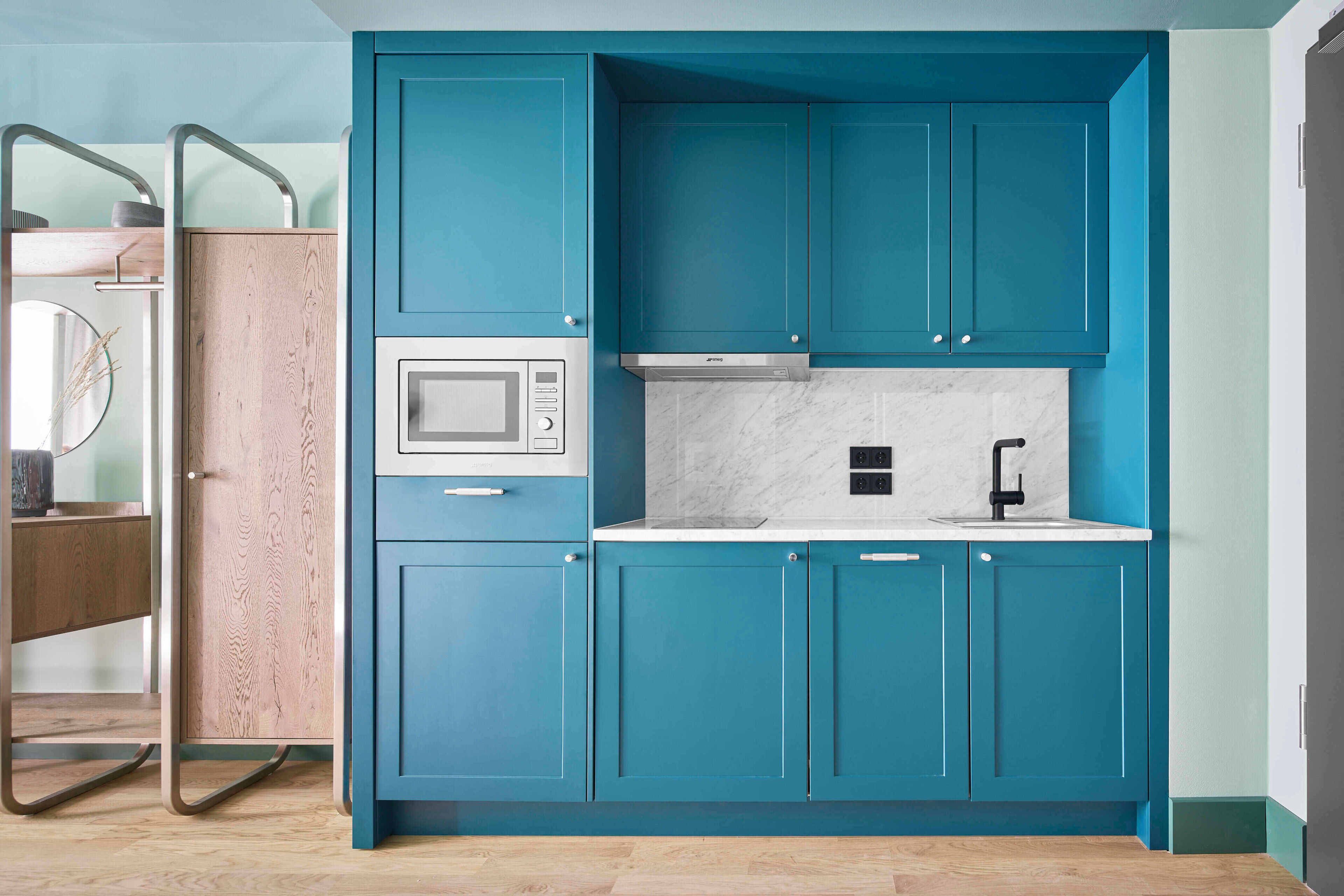 A compact kitchen with teal cabinetry, a microwave, a sink, and a wooden cabinet.