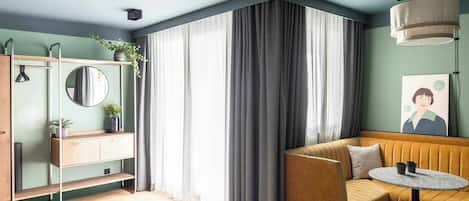 A modern hotel room with a bed, a sofa, a small table, a mirror, a shelf with plants, and a large window with curtains.