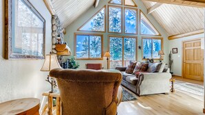 A cozy loft-style living room with a high ceiling, large windows, and a view of snowy trees. - Private Hot Tub in the Aspens - Steam Shower, Log Fireplace, Mtn Views (Alma)