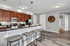 A modern kitchen with wooden cabinets, a stainless steel microwave, and a refrigerator. There are bar stools and pendant lights.