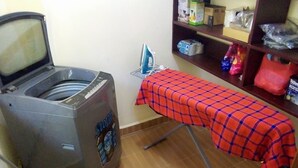 Un'area lavanderia con lavatrice, un asse da stiro con coperta a quadri e scaffali con diversi oggetti.