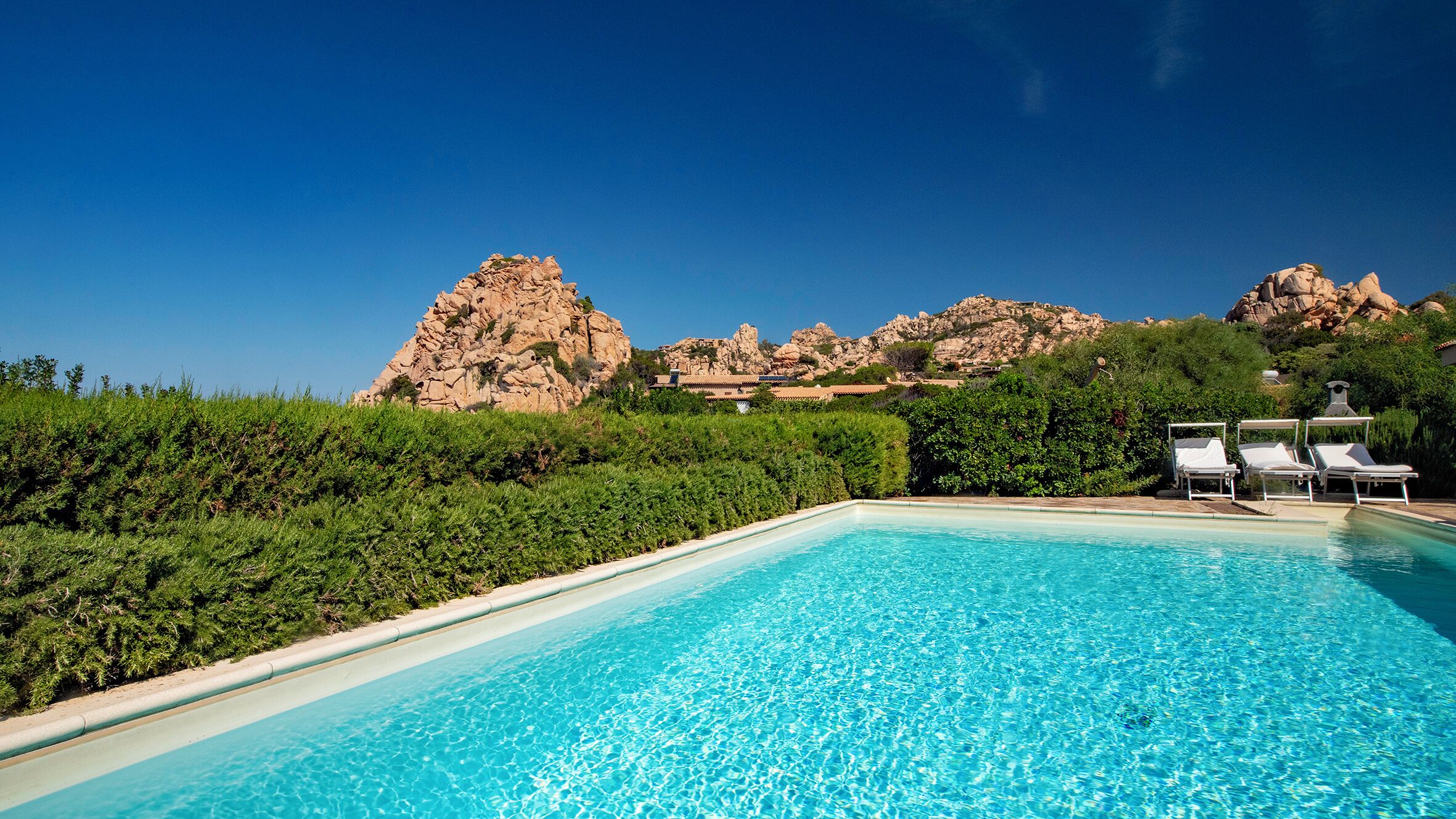 A swimming pool with clear blue water, surrounded by lush greenery and rocky hills in the background.