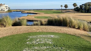 Un parcours de golf avec un plan d’eau, des palmiers et un grand bâtiment en arrière-plan.