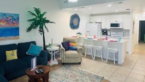 Una sala de estar con sofá, sillón y una pequeña mesa de centro. Se observa una zona de cocina con armarios blancos y un microondas. Hay un ventilador de techo, y un cuadro con una escena de playa en la pared.