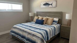 Un dormitorio con una cama grande, dos mesitas de noche, un cuadro colgado en la pared con un tiburón y una ventana con persianas.