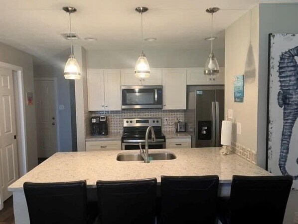 A kitchen with a large island, stainless steel appliances, pendant lighting, and a refrigerator.