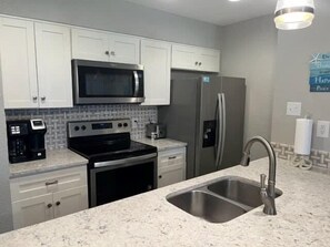A kitchen with white cabinets, a stainless steel refrigerator, a microwave, and a double sink. - Island Dunes Condo's-#1109-One Block from Beach. (Port Aransas)