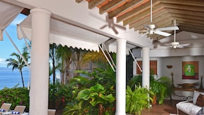 A covered outdoor seating area with white furniture, a ceiling fan, and a view of the ocean.