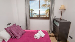 Quarto de hotel com cama, escrivaninha, luminária e janela com vista para a praia.