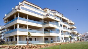 A multi-story residential building with balconies, a stone retaining wall, and a grassy area in front. - Marina del Castillo 321 (Manilva)