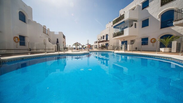 A swimming pool with clear blue water, surrounded by white buildings with balconies and windows. - Mikonos Playa apto 19 (Manilva)