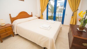 A neatly made bed with white linens and towels, a wooden nightstand with a lamp, and a wooden dresser with a plant. - Mikonos Playa apto 19 (Manilva)