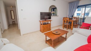 A living room with a TV, wooden cabinet, dining table, and chairs. - Mikonos Playa apto 19 (Manilva)
