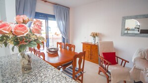 A dining area with a table, chairs, a vase of flowers, and a view through a window with curtains. - Mikonos Playa apto 19 (Manilva)