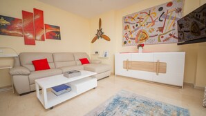 A living room with a beige sofa, a white coffee table with a book, a ceiling fan, and abstract wall art. - Mikonos Playa 14 (Manilva)