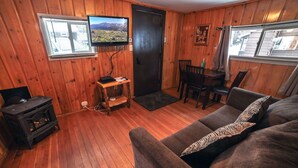 A cozy cabin interior with a wooden floor, a sofa, a dining table with chairs, a television mounted on the wall, and a wood stove. - Cabin offers a rustic and cozy getaway (Estes Park)
