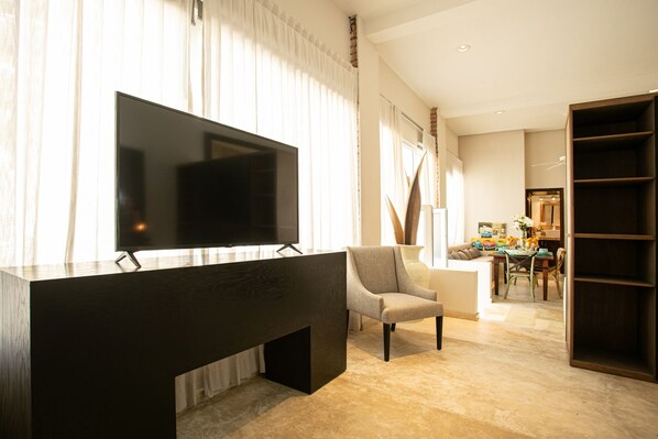 A modern living room with a large flat-screen TV, a dark wooden console table, a grey armchair, and a dining area with a table and chairs.
