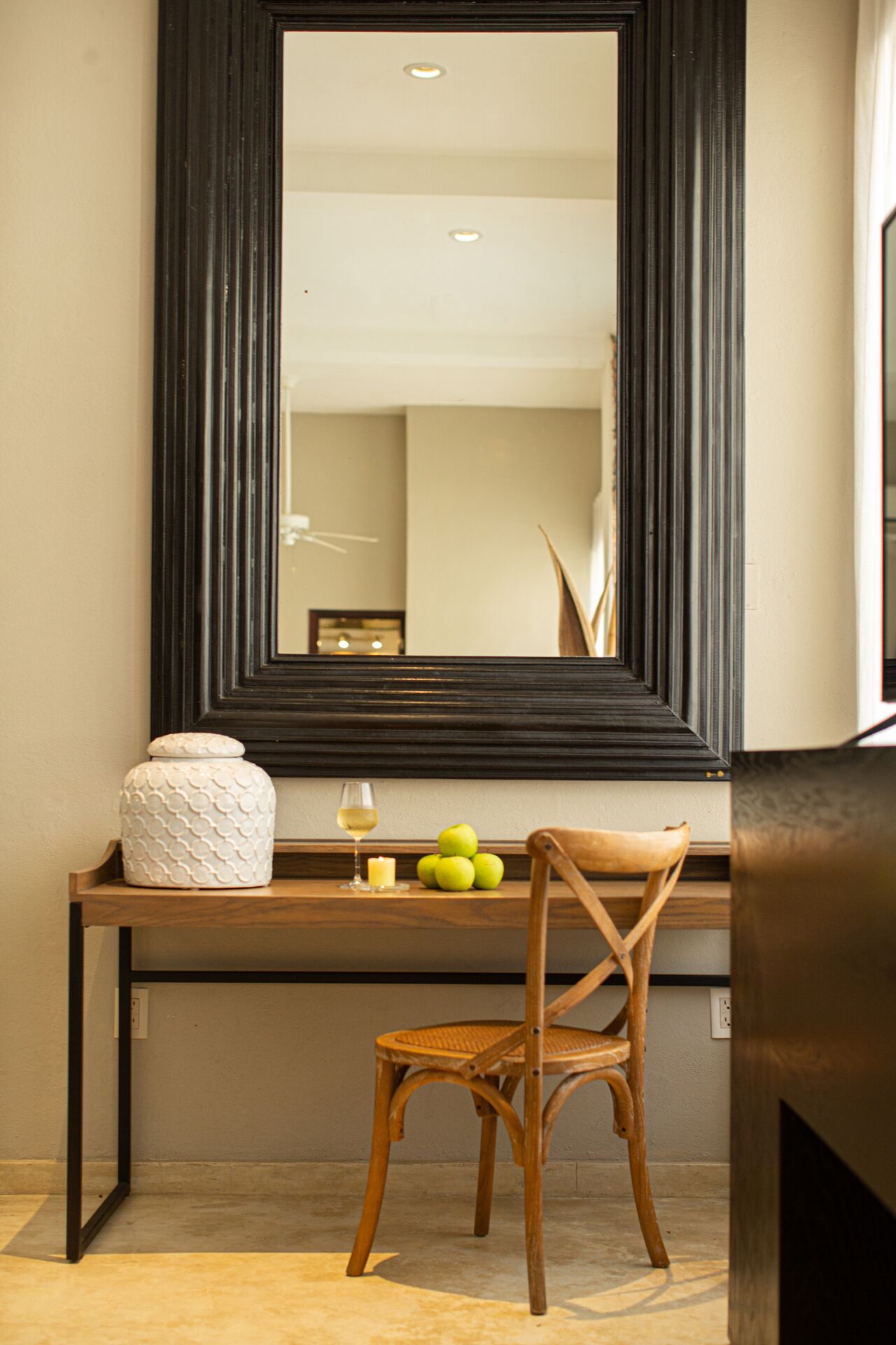 A room with a large mirror, a wooden chair, a small table with a white vase, a candle, and a glass of wine.