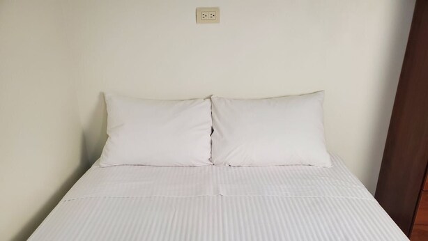 Una cama individual con colchón y almohadas blancas, una pared lisa y un armario de madera a la derecha.