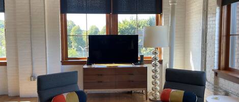 Una sala de estar moderna con un televisor de pantalla plana sobre un mueble de madera, dos sillones grises con cojines coloridos, una mesa de centro de vidrio y una lámpara blanca.